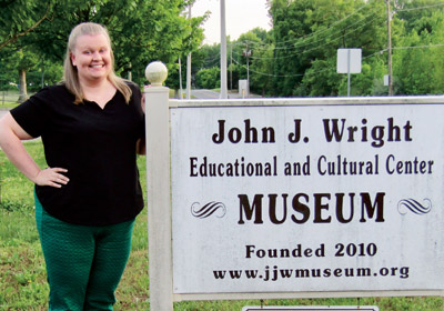 Woman stands next to a sign that reads John J. Wright Educational and Cultural Center Museum Founded 2010 www.jjwmuseum.org