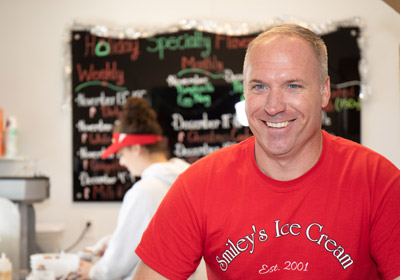 Man in red shirt smiling in foreground. Young woman in background scooping ice cream.|