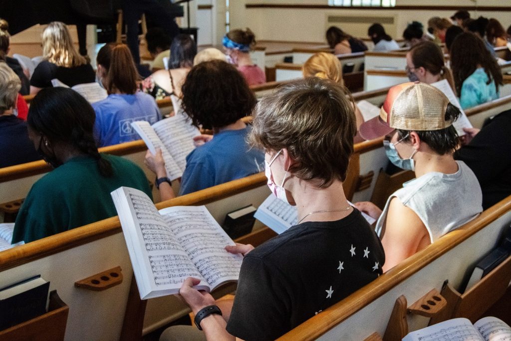 Two males hold song books while sitting in pews for a choir rehearsal