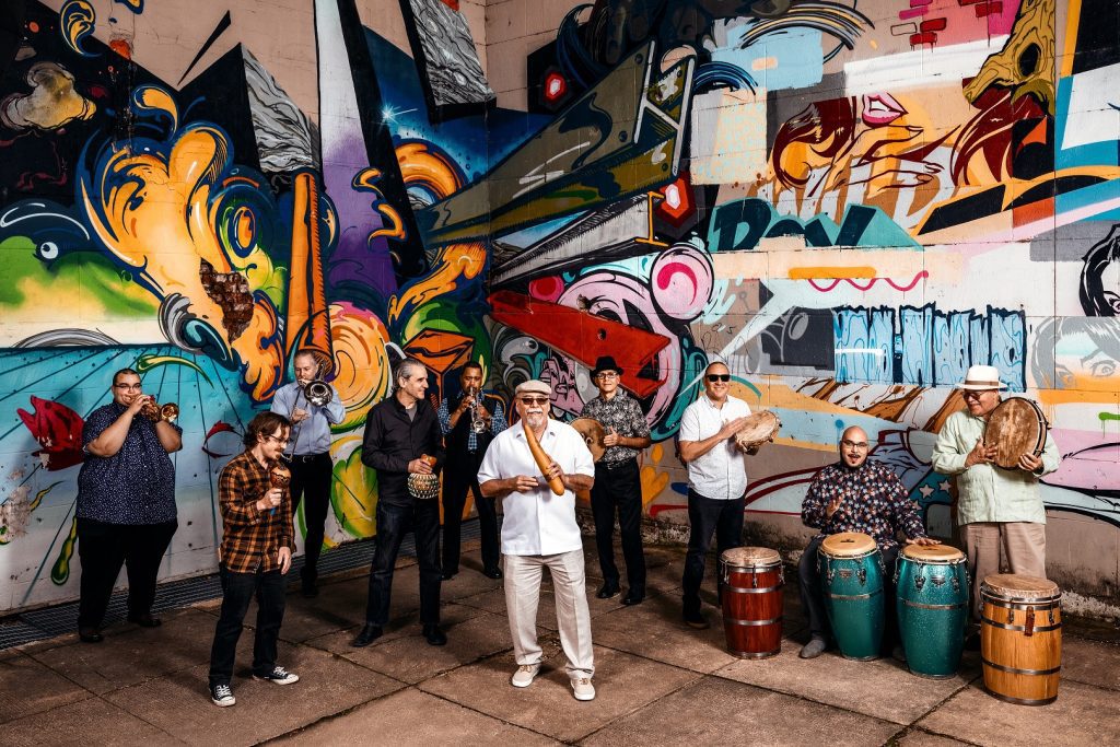 A 13-piece band stands in front of a colorful mural