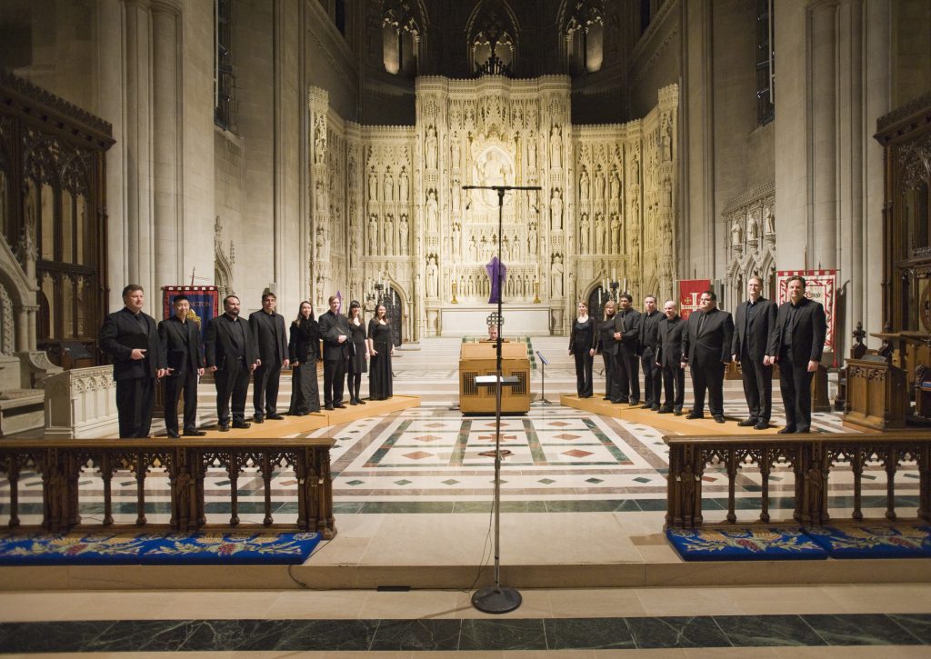 Choir members dressed in all black stand in a church. A microphone is centered around them
