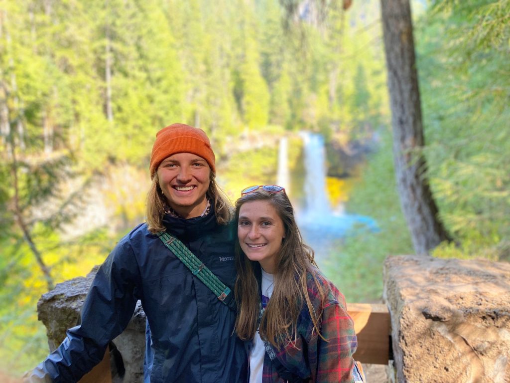 A young male and female stand side by side smiling for a posed photo outdoors. A waterfall can be seen in the background