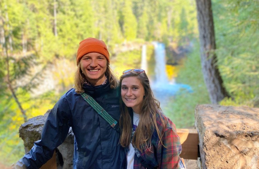 A young male and female stand side by side smiling for a posed photo outdoors. A waterfall can be seen in the background