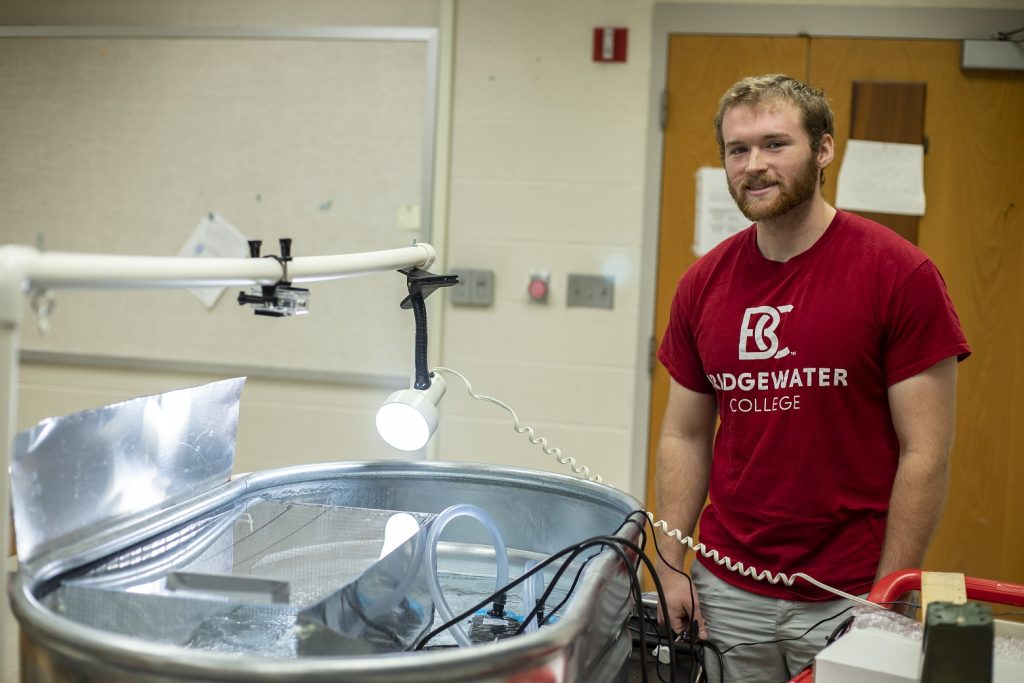 A male student stands by a fish tank he made to test water flow and current preference of longnose dace, a small freshwater fish. He is smiling and wearing a Bridgewater College t-shirt.