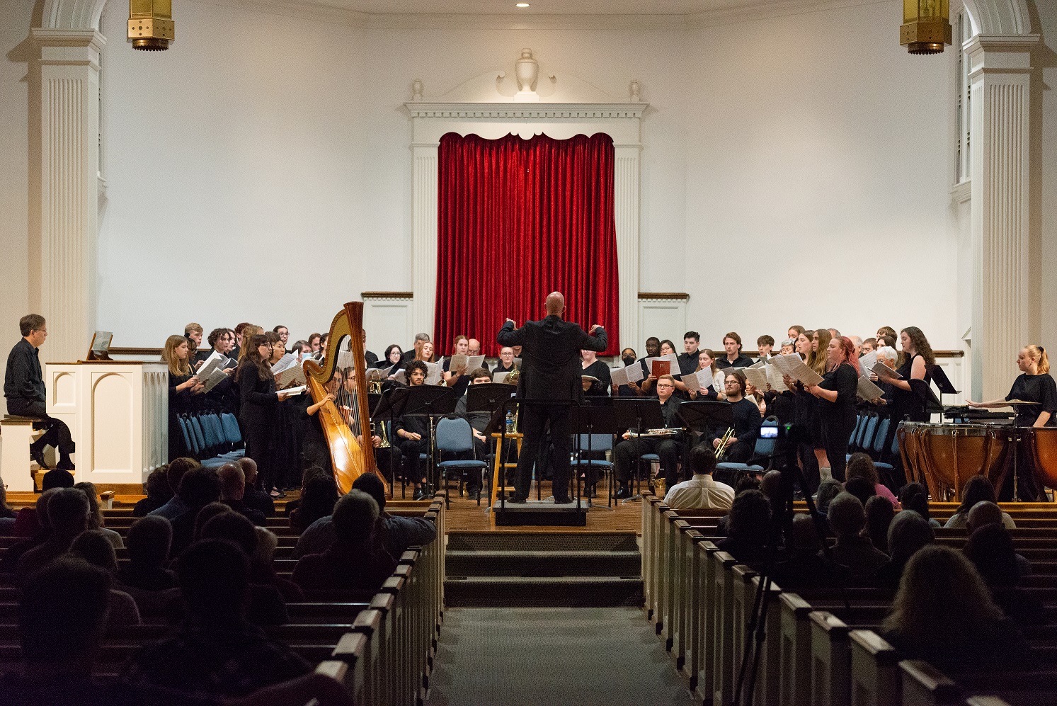 A music conductor stands at center stage with his arm raised and back to the audience. Members of the choir stand before him, facing the audience. The audience is seated in pews.