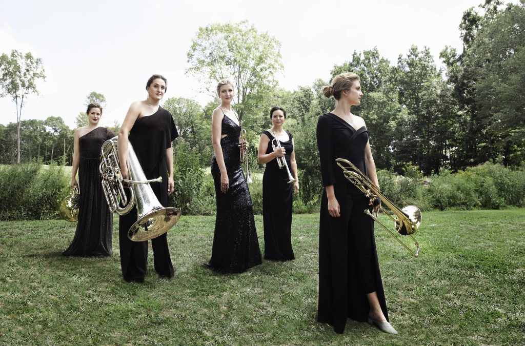 Five women dressed in black gowns standing outside on green grass holding brass instruments.
