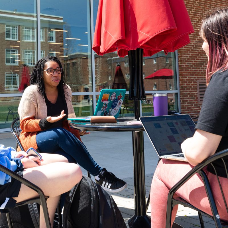 Students sitting at a table with an umbrella talking outside the Forrer Learning Commons