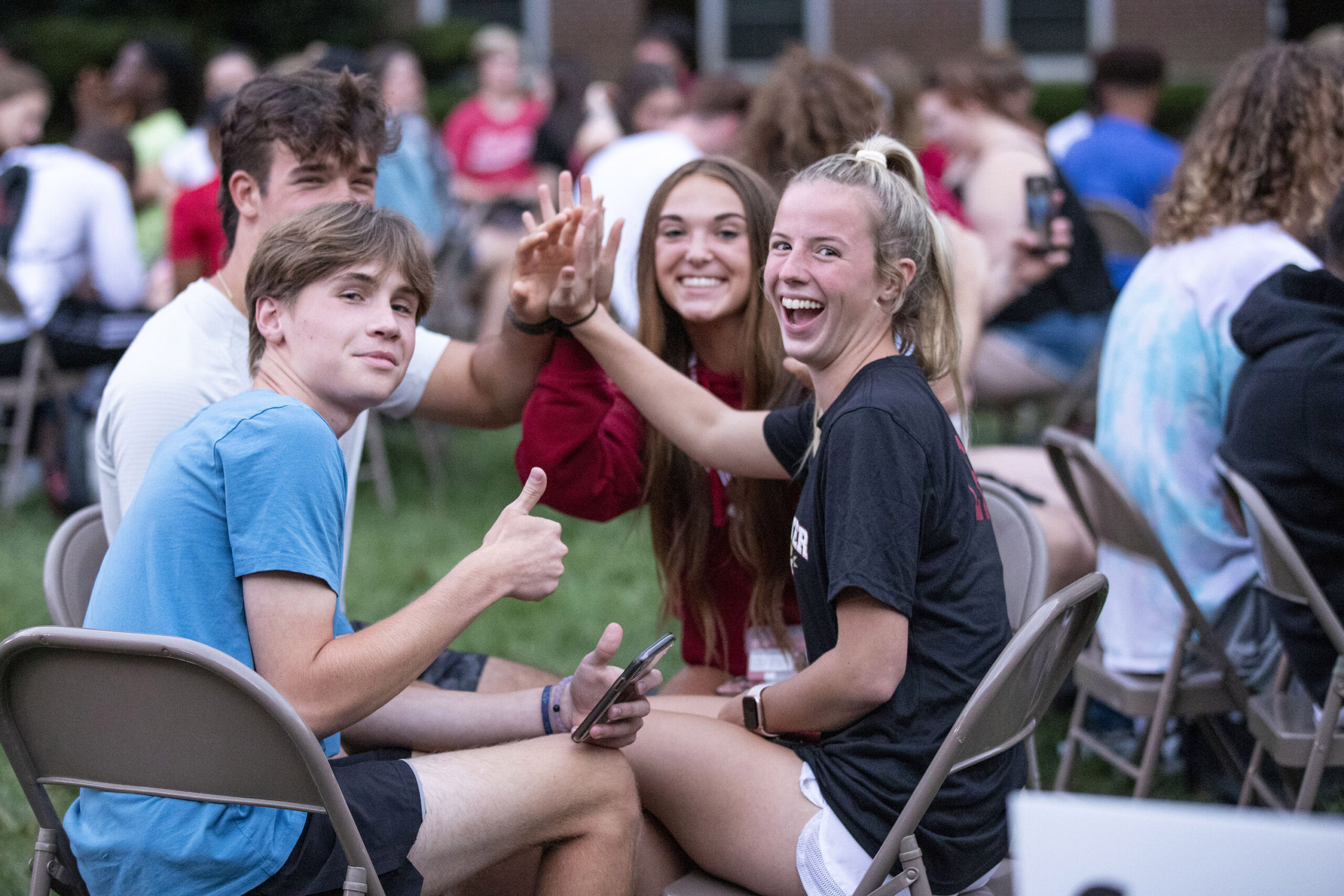 Group of boy and girls sitting in folding chairs forming a circle and high fiving in the middle. One boy on the left is giving a thumbs up to the camera.