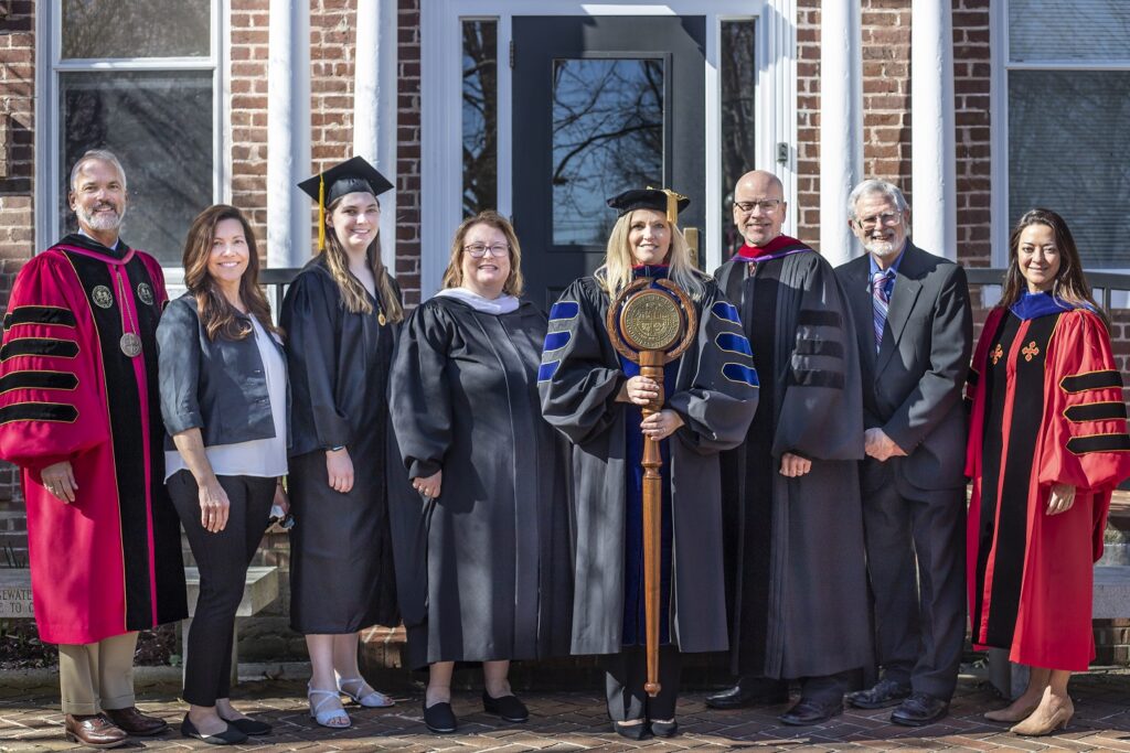 Eight men and women stand in front of a brick building smiling and wearing academic regalia.