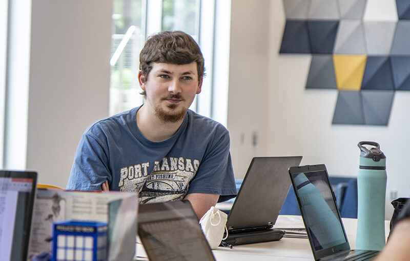 Student sitting at a table with several laptops on the table