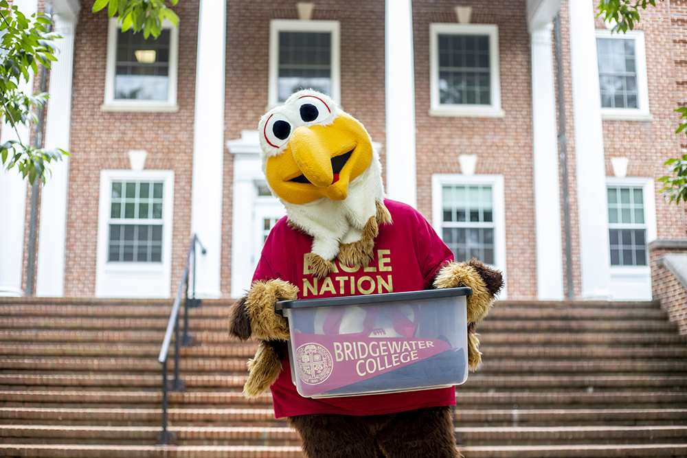 Bridgewater College Mascot Ernie holding a bin of stuff in front of brick building