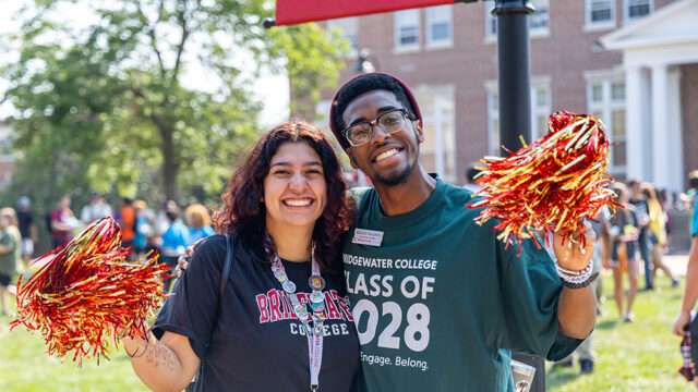 Two students standing next to each other smiling and waving gold and crimson pom-poms