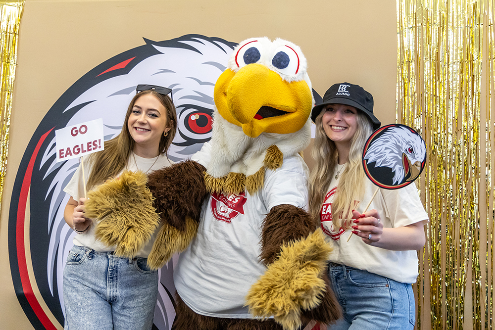 Bridgewater College alumni pose with the BC Eagle mascot at a festive event. Two women wearing BC Alumni shirts and accessories smile while holding signs that say 'Go Eagles!' and feature the eagle logo. The background is decorated with gold streamers and a large eagle graphic.