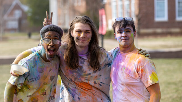 Three students posing for a photo during the Holi event covered in bright colored chalk