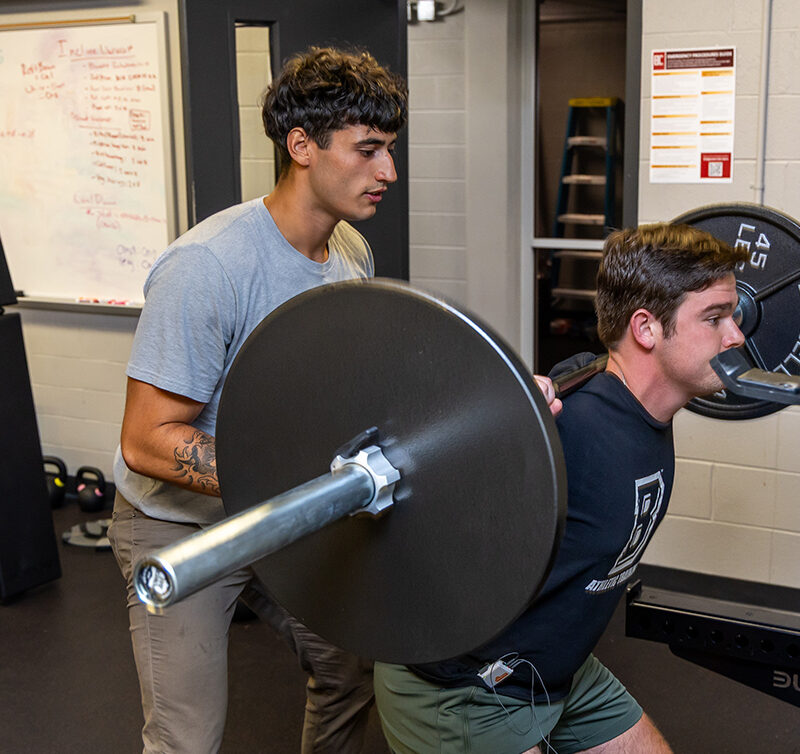 Student squating at squat rack with another student spotting