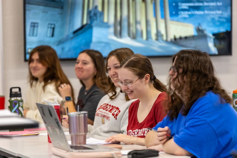 Five students sitting next to each other at a row of desks and laughing