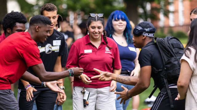 Two students play rock paper scissors while several onlooking students cheer them on